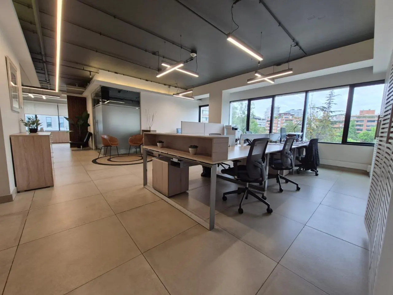Bright, modern office space with desks, chairs, and large windows overlooking a city. Neutral color palette with wood accents.