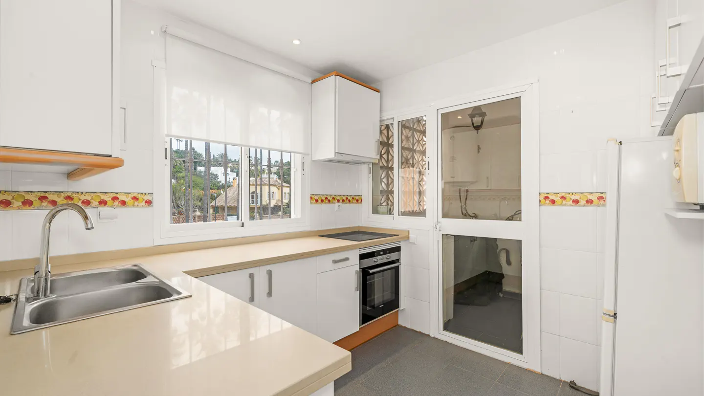 Bright kitchen with white cabinets, beige countertops, and stainless steel sink. A window with bars overlooks a building. A glass door leads to another room.