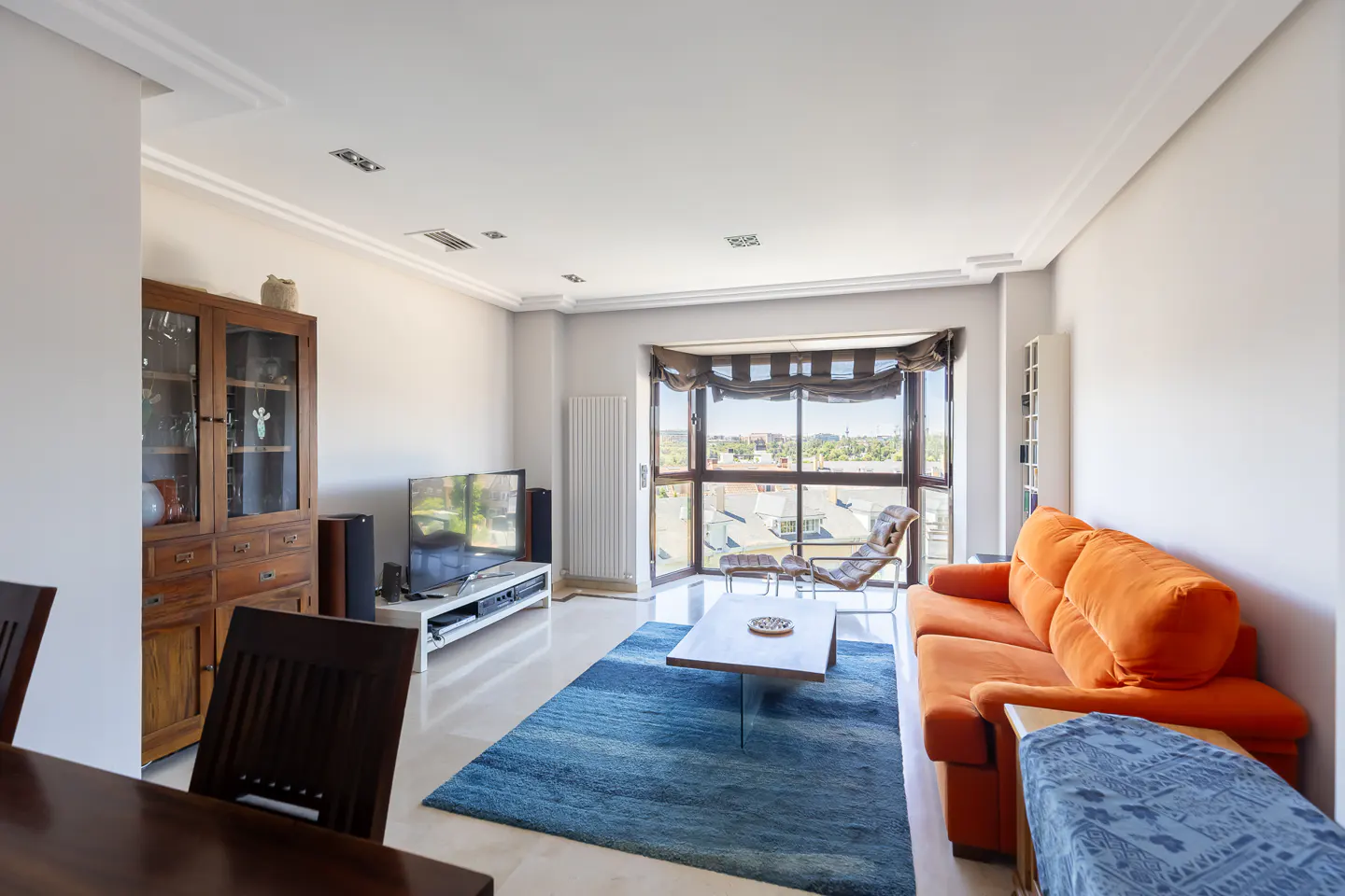Bright living room with an orange sofa, blue area rug, wooden coffee table, and a large window with a city view.