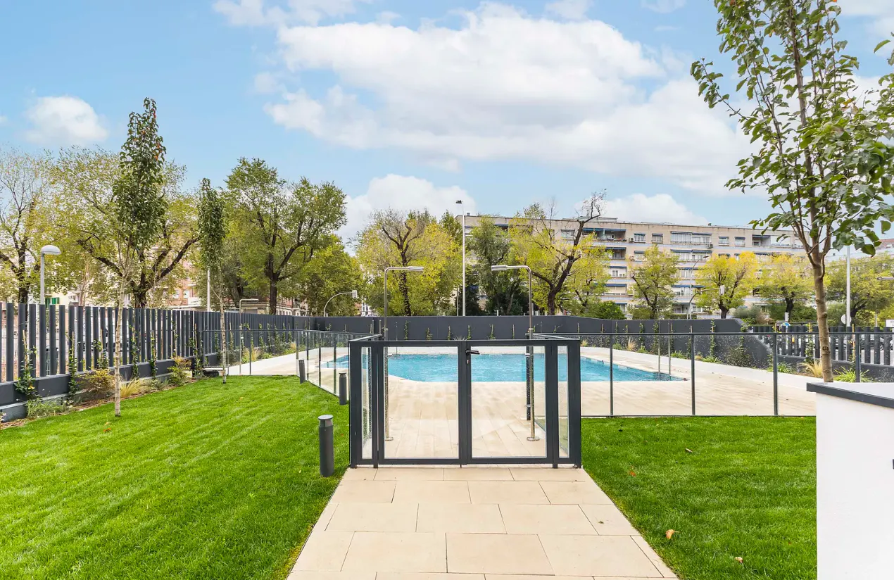 A fenced-in outdoor swimming pool surrounded by a green lawn, paved walkway, and trees under a blue sky with clouds.
