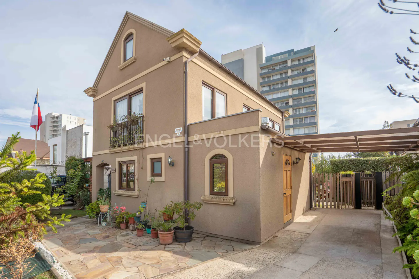 Exterior view of a two-story tan house with a balcony, arched windows, and a covered driveway. A Chilean flag is visible in the background.