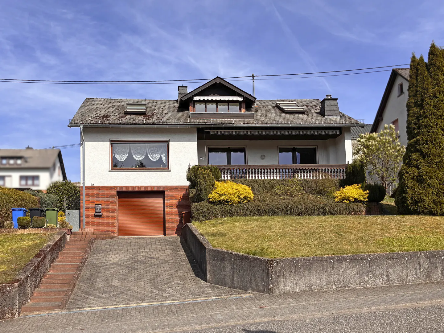 Two-story house with a brown garage door, white walls, and a gray roof under a blue sky. A lawn and shrubs are in front.