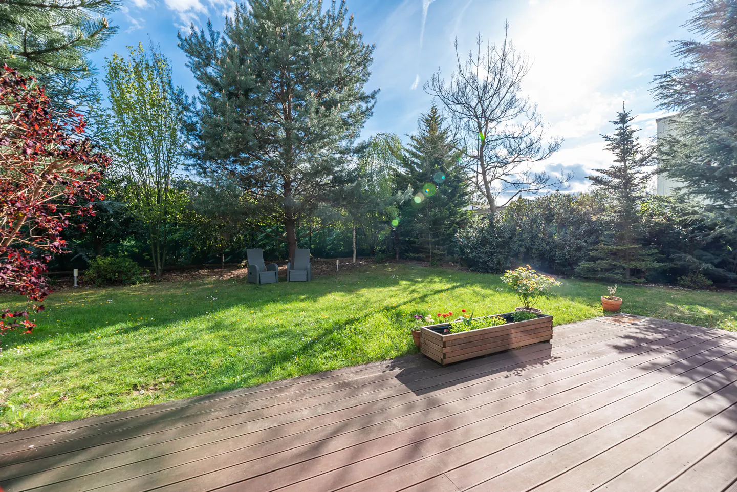 Wooden deck overlooking a green lawn with trees and two chairs in the background. A wooden planter box with flowers sits on the deck.
