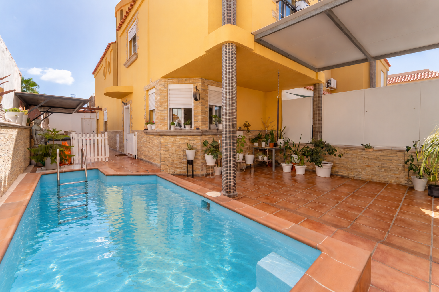 A yellow house with a swimming pool, tiled patio, and various potted plants under a covered outdoor area. [AI-generated text] [AI-modified]