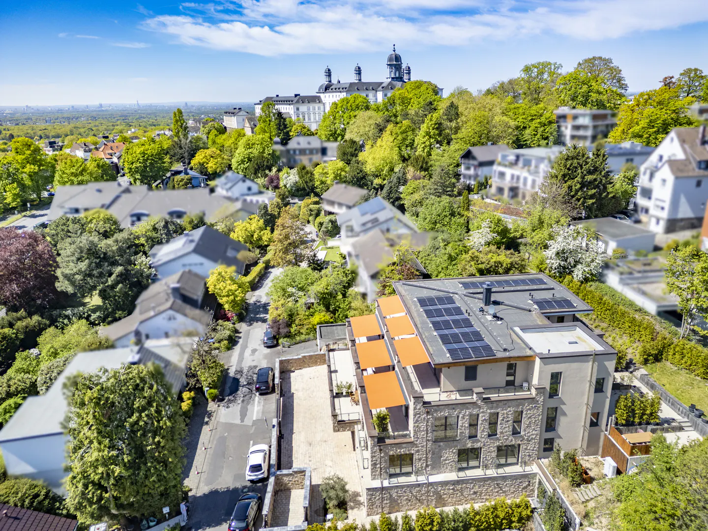 Aerial view of a stone building with solar panels and orange awnings, surrounded by trees and houses. A church is visible in the background.