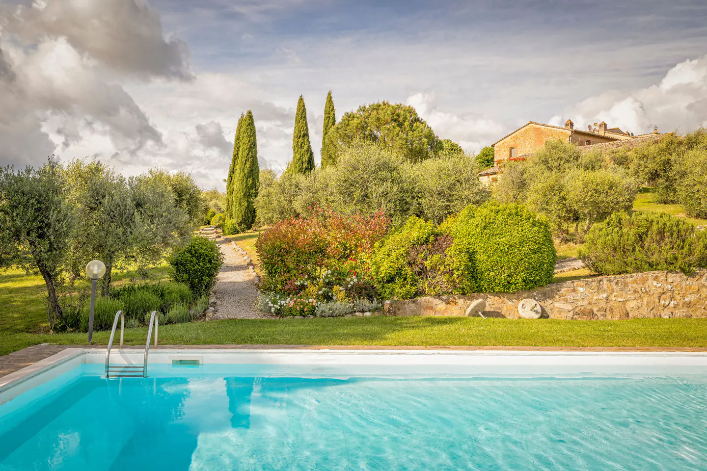 A bright blue swimming pool in the foreground with a gravel path leading to a stone villa surrounded by trees and shrubs.