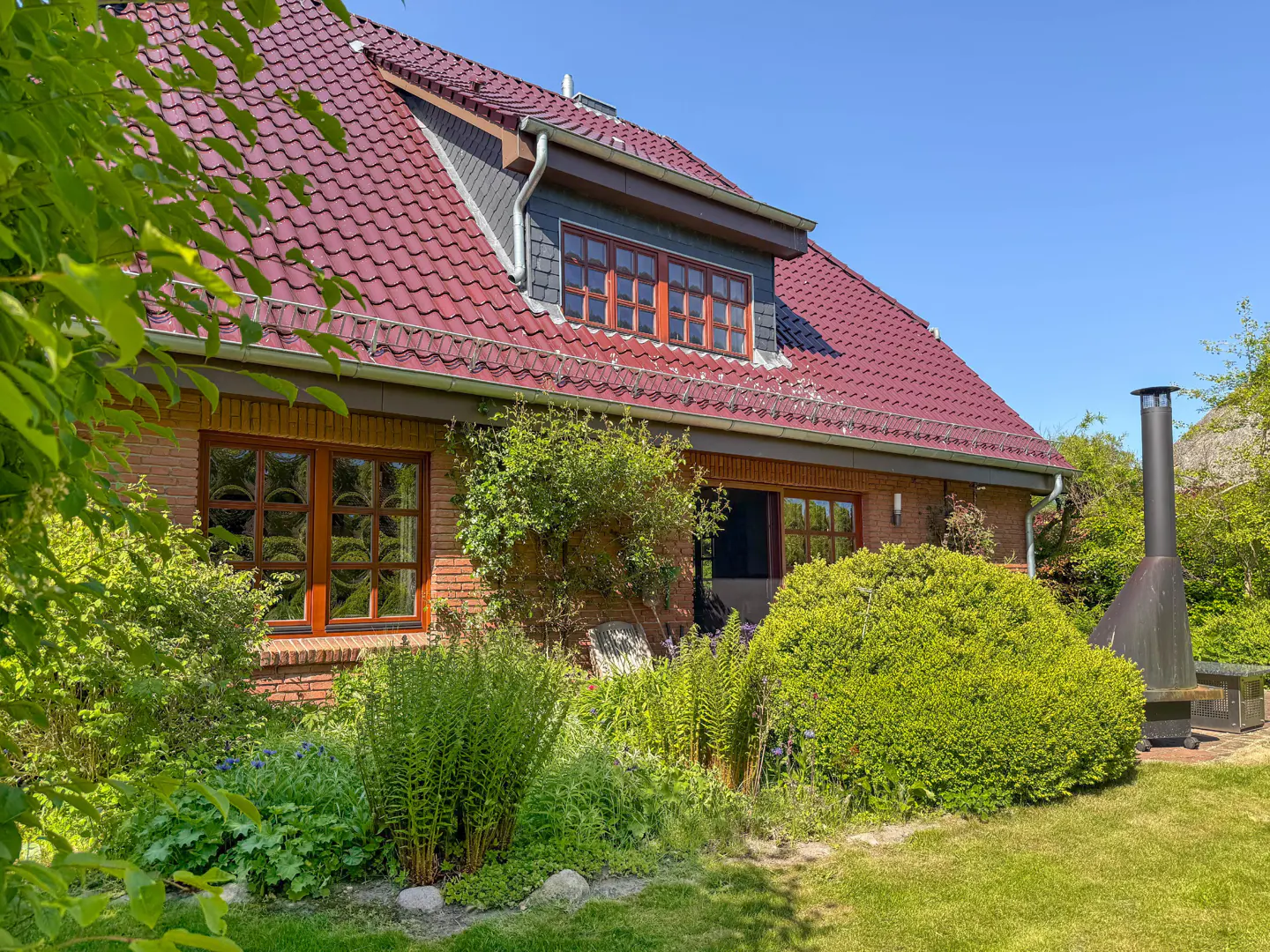 Brick house with a red tile roof, brown window frames, and green bushes in the yard. A black chimney stands on the right.