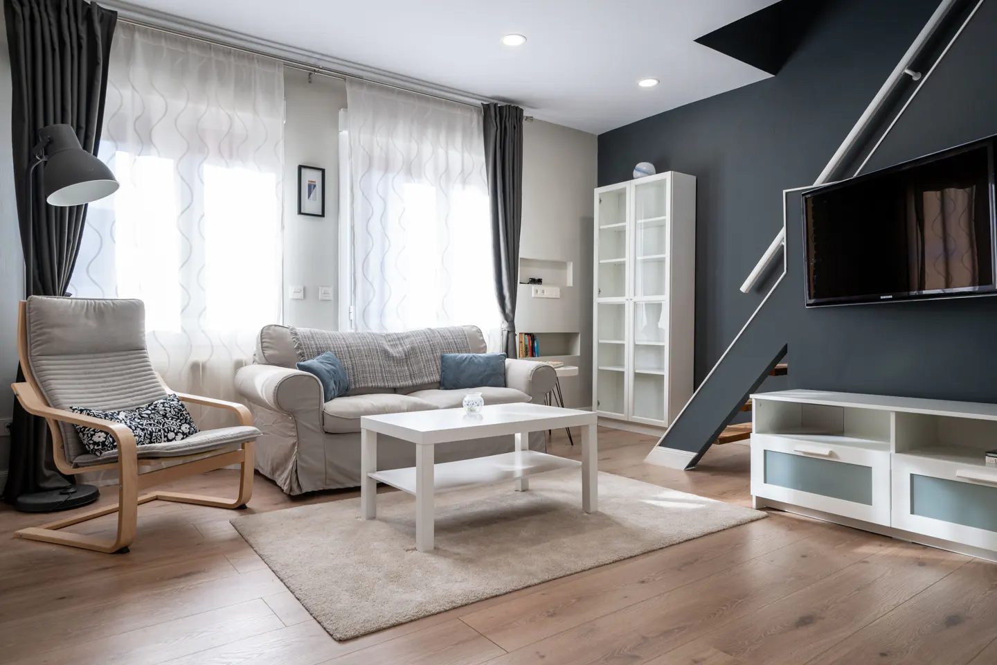 Living room with a gray sofa, chair, and white table on a light rug. A TV is mounted on a dark blue wall with white shelves and stairs.