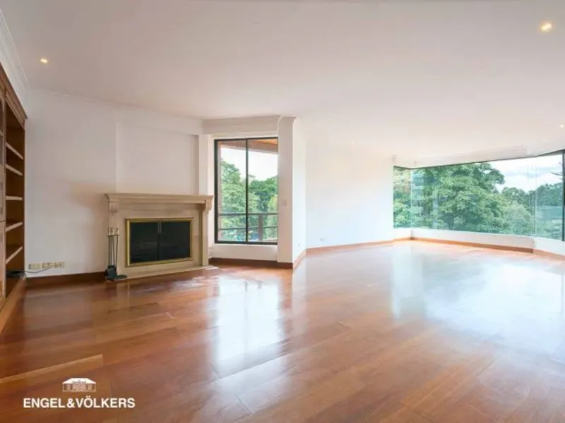 Bright, empty living room with hardwood floors, white walls, a fireplace, and large windows with a view of green trees.