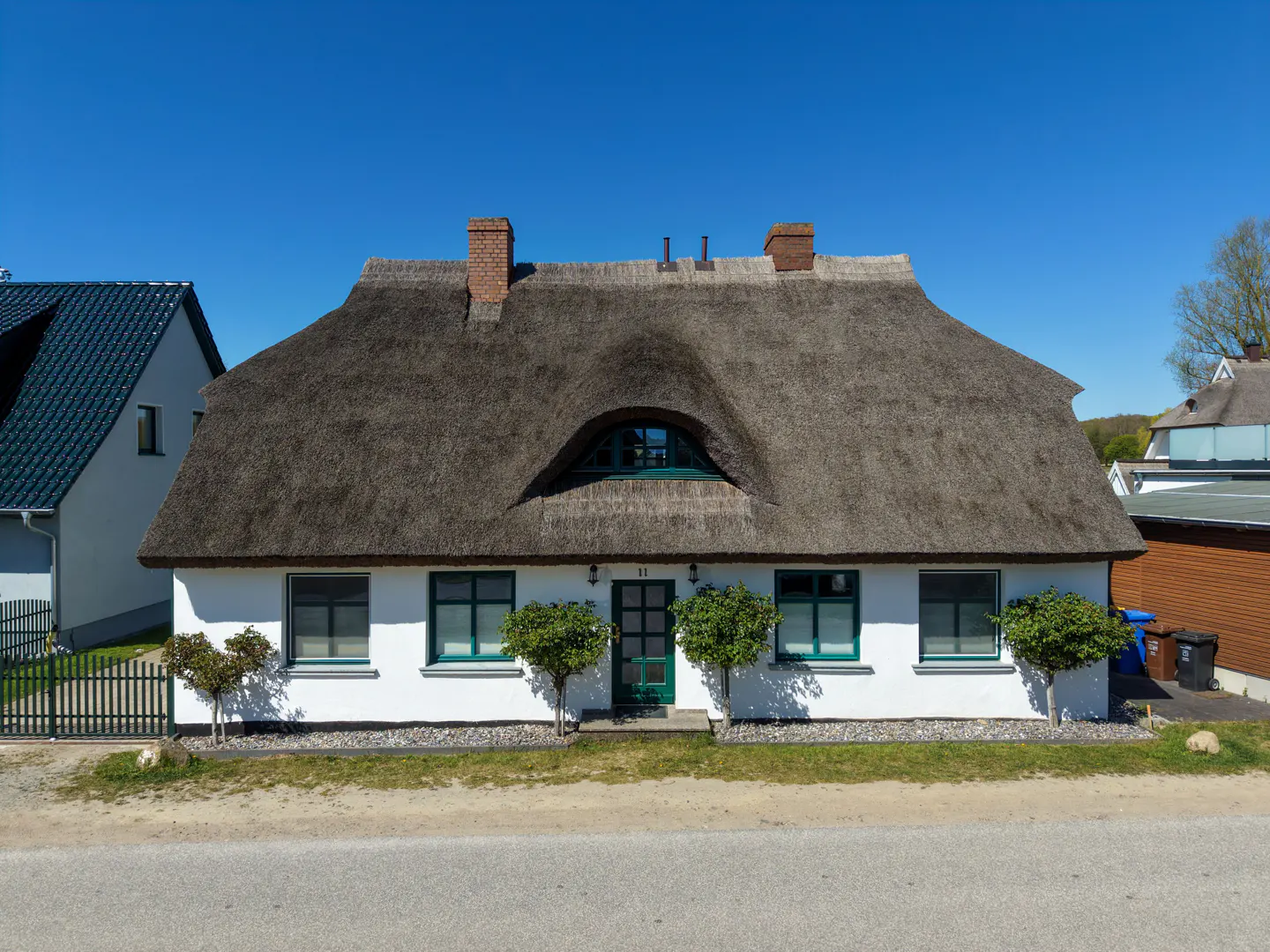 Charming white house with a thatched roof, green trim, and manicured trees under a clear blue sky.