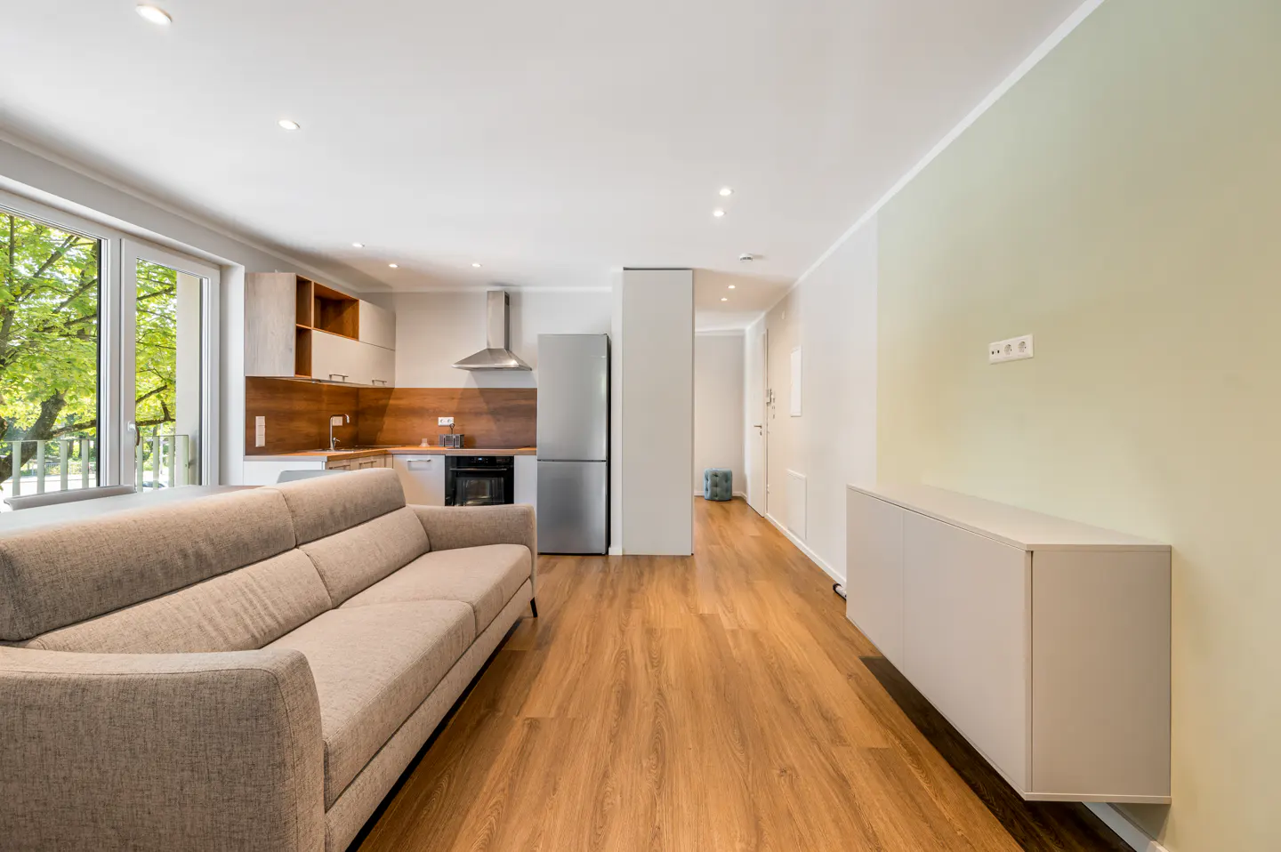 Bright apartment interior with a beige sofa, wood floors, and a kitchen with stainless steel appliances. A large window shows green trees outside.