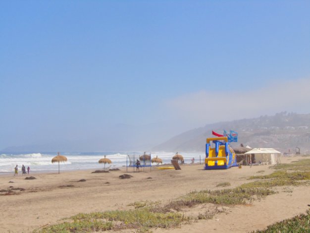 Sandy beach scene with blue sky, ocean waves, and distant hills. Features straw umbrellas, a trampoline, and a blue and yellow inflatable slide.