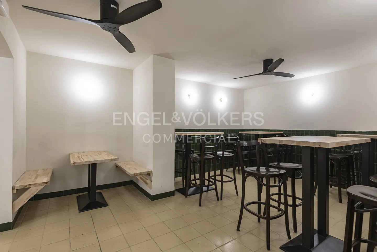 Interior of a commercial space with tables, chairs, and ceiling fans. Walls are white, and the floor is tiled.