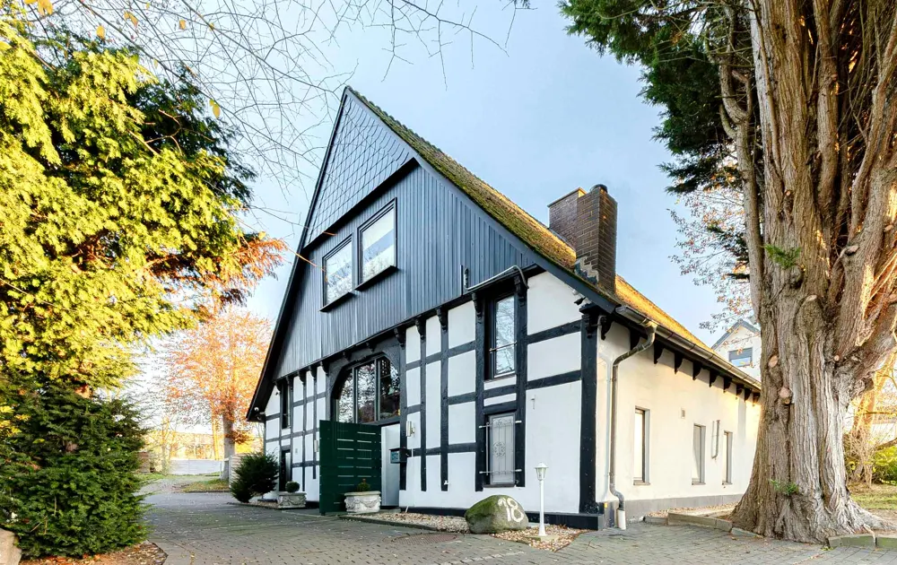 Exterior view of a two-story black and white Tudor-style house with a brick chimney and a large tree on the right.