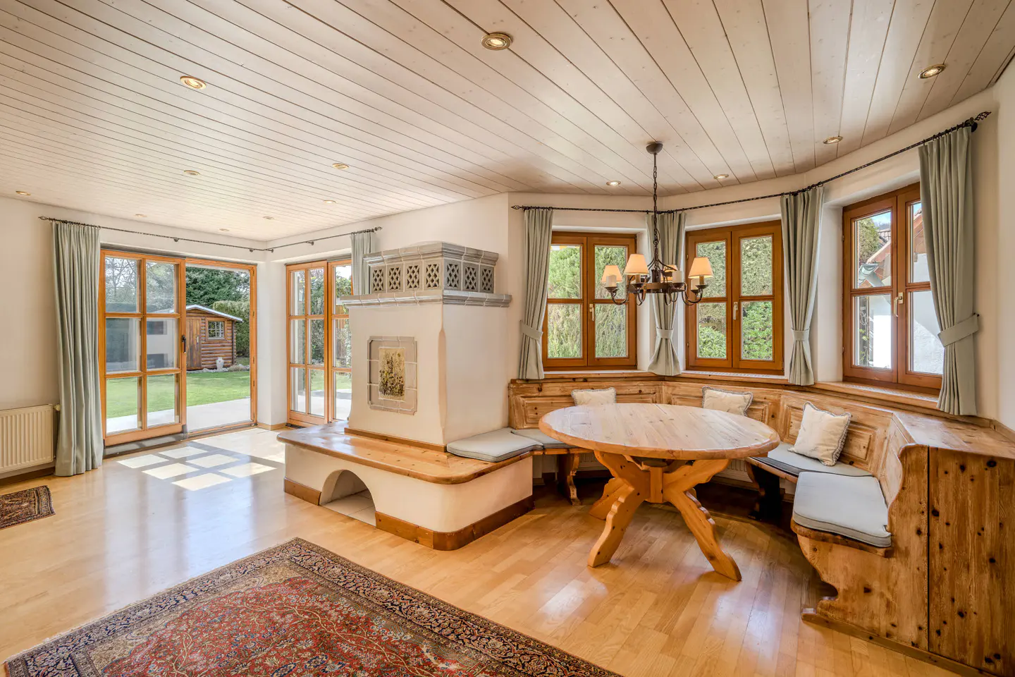 Bright dining room with wood floors, a round table, and a built-in bench. A tiled stove sits near open doors to a green yard.