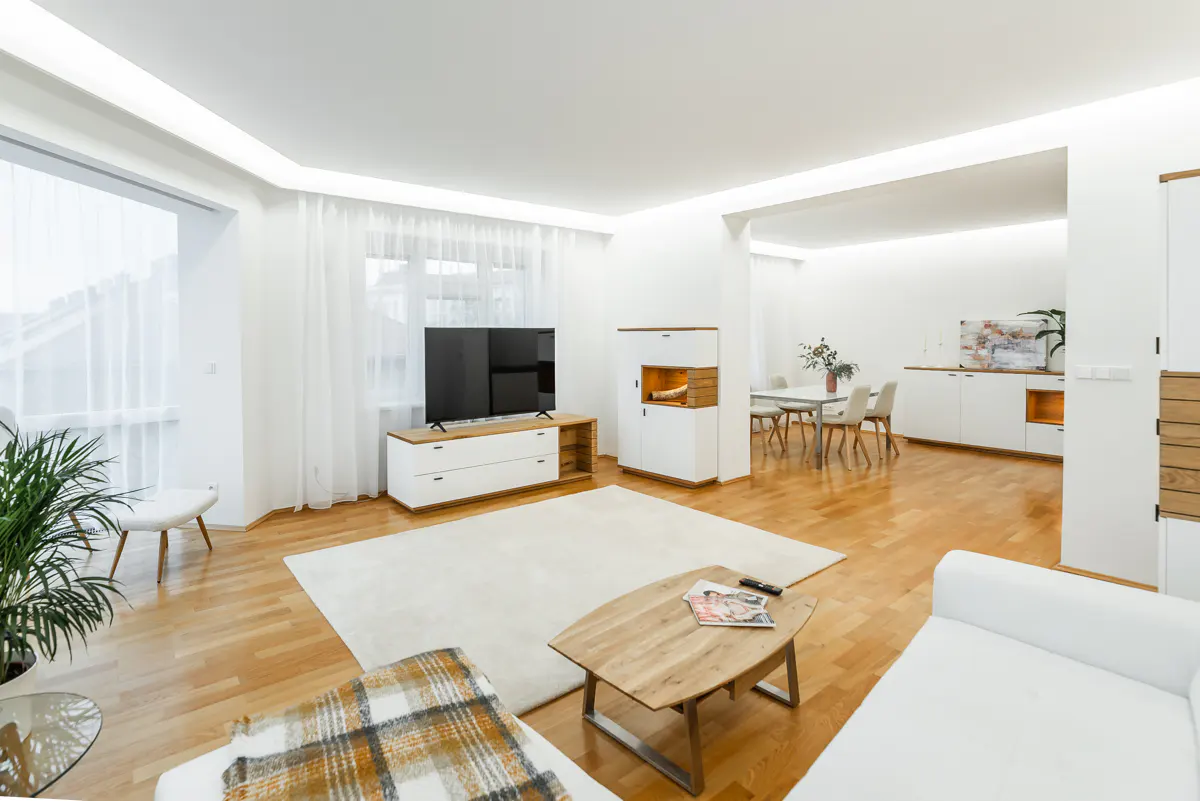 Bright, modern living room with hardwood floors, white walls, and recessed lighting. A TV, fireplace, dining table, and white sofa furnish the space.