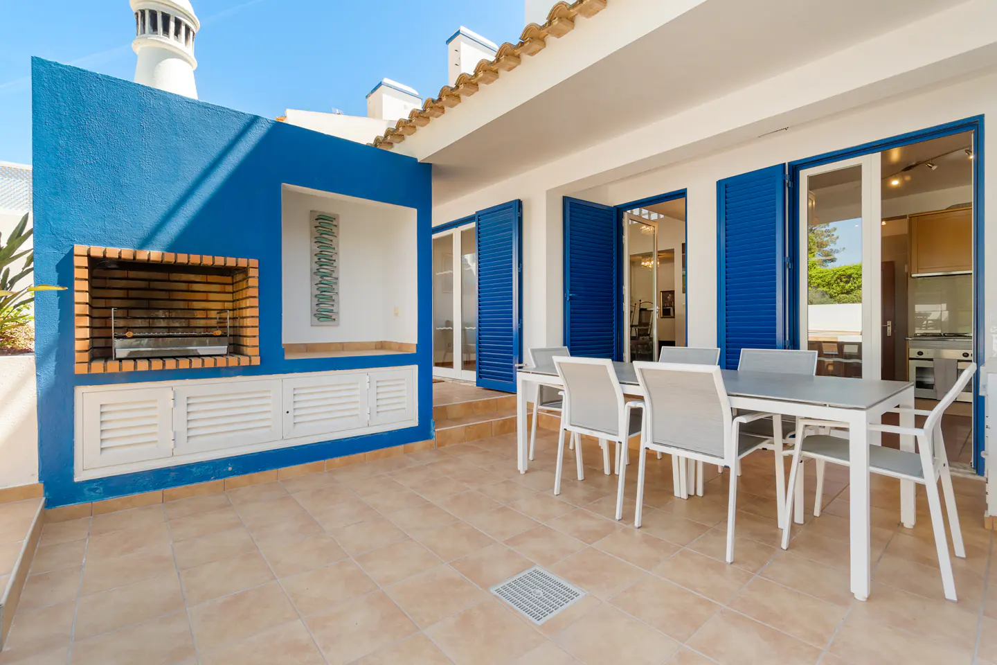 Outdoor patio with a blue wall, brick grill, and white cabinets. A table and chairs sit near open blue shuttered doors.