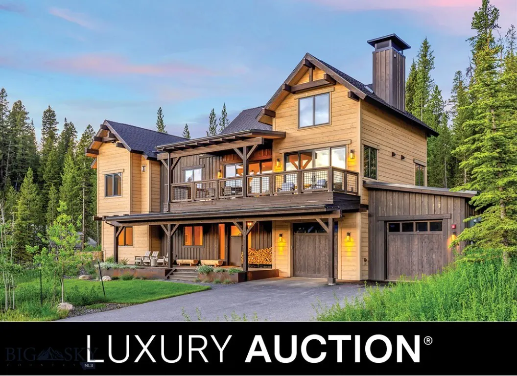 A two-story luxury home with wood siding, a dark roof, and a large porch, surrounded by green trees.