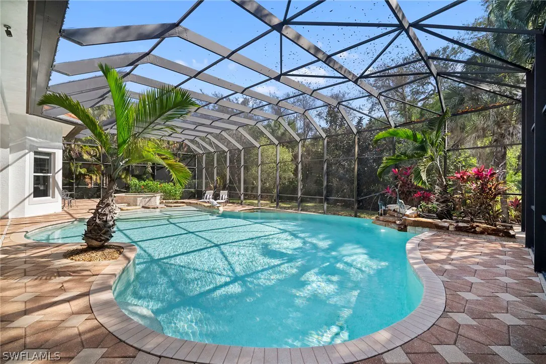 A screened-in pool with turquoise water, surrounded by brick pavers and lush greenery. A palm tree grows near the pool's edge.