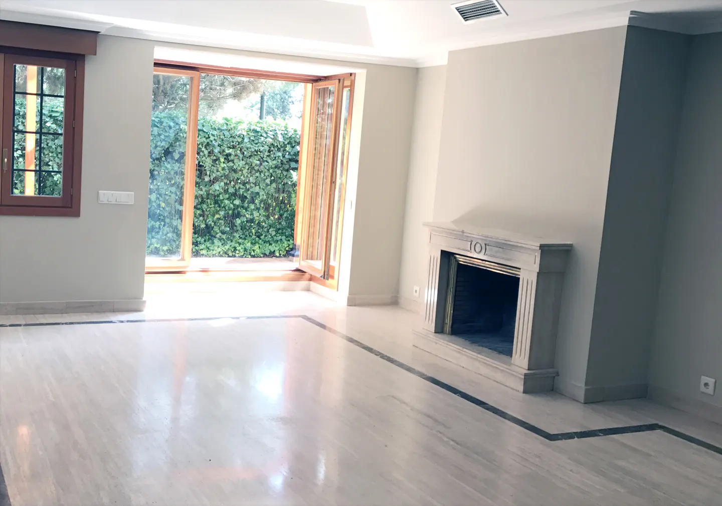 Bright living room with light marble floors, a stone fireplace, and large glass doors opening to a lush green garden.