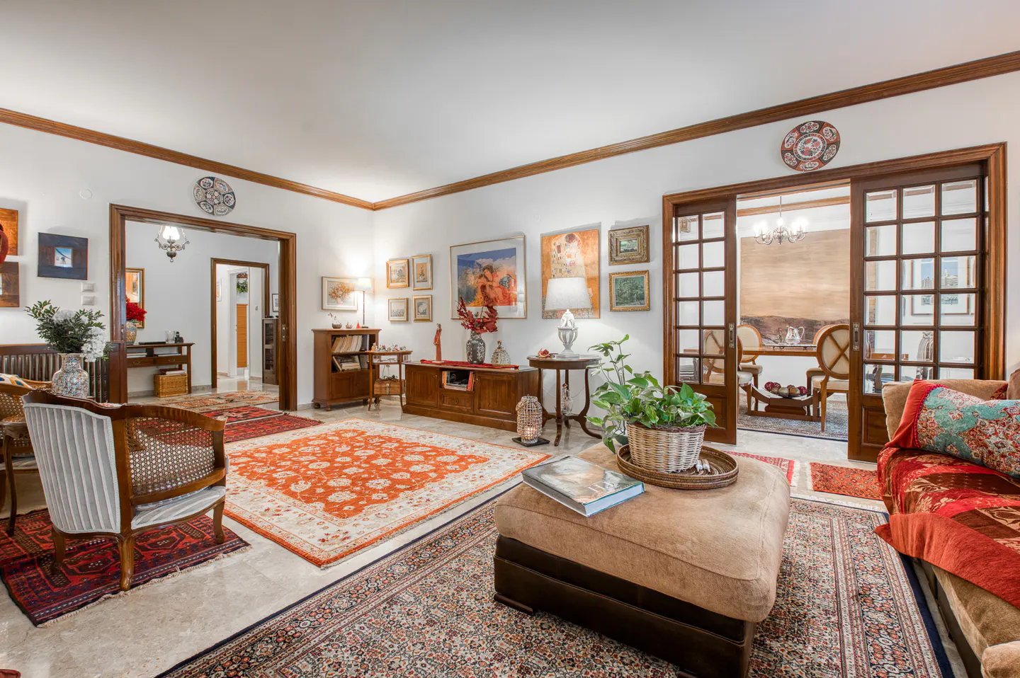 A living room with white walls, wood trim, and multiple rugs. An ottoman sits in the foreground, with a view into the dining room.