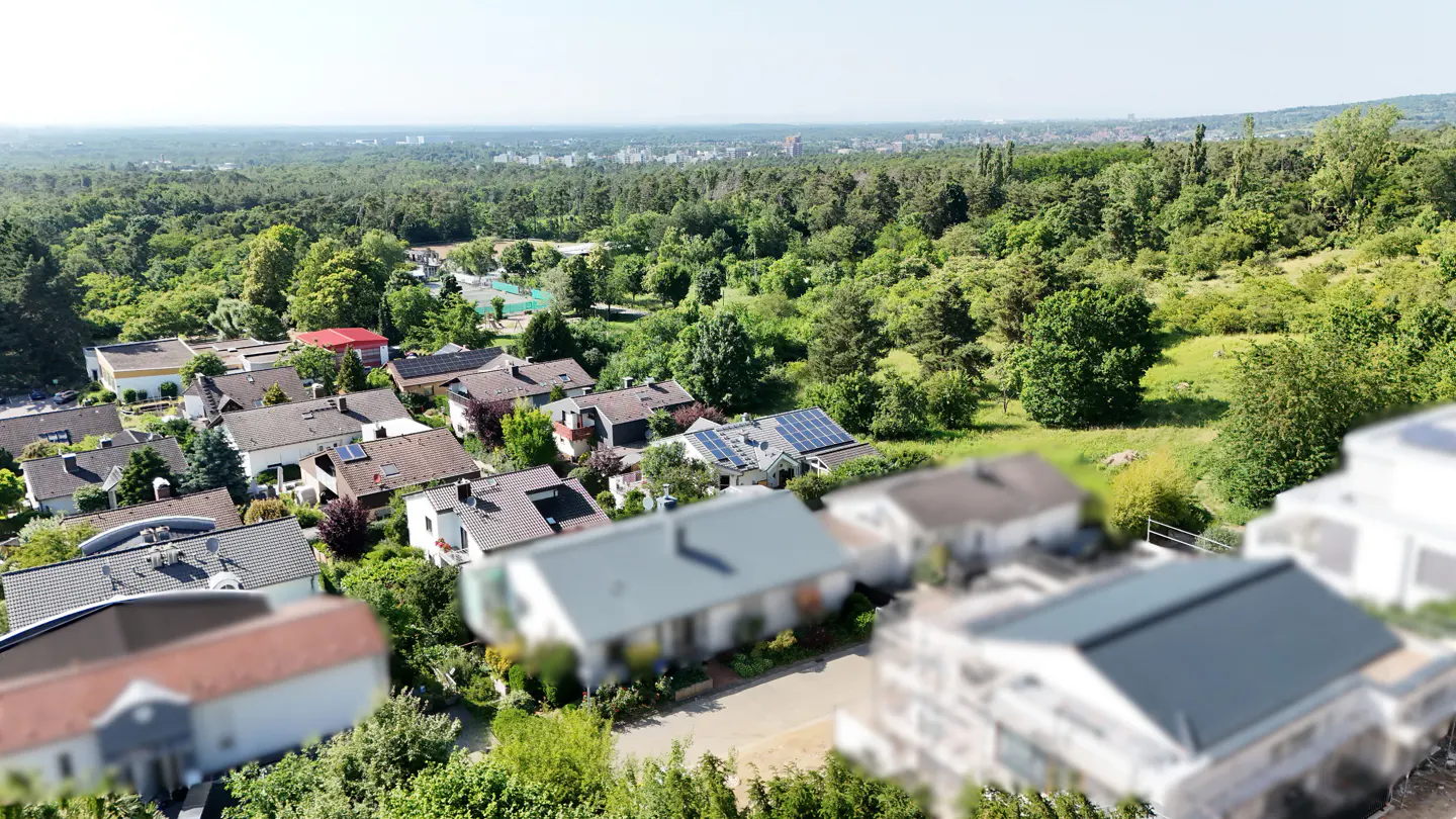 Aerial view of a residential area with houses, green trees, and a forest in the background. Some houses have solar panels on their roofs.