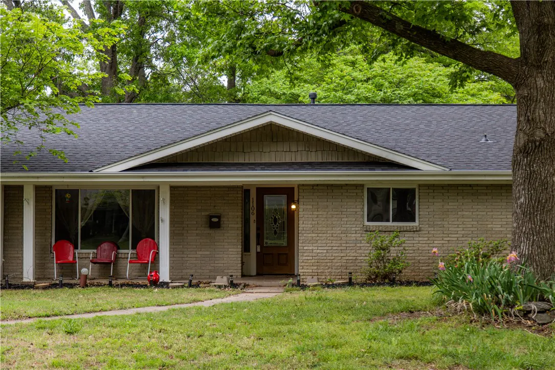 Front view of a one-story brick house with a dark roof and green lawn. Three red chairs sit on the porch. A tree is on the right.