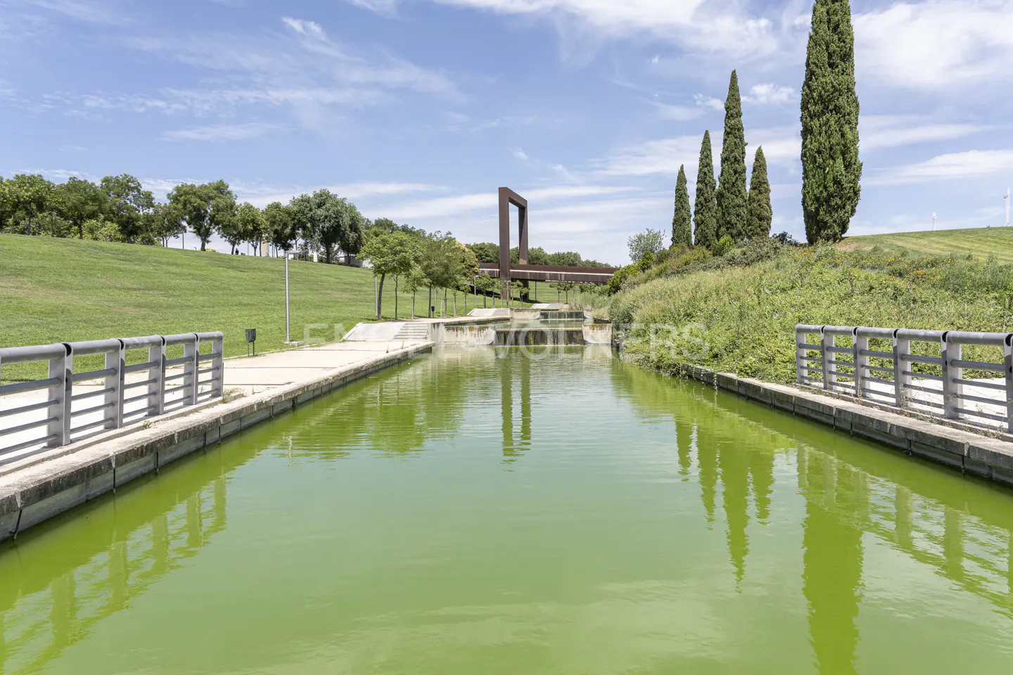Green canal with metal railings on each side, leading to a modern sculpture and trees under a blue sky.