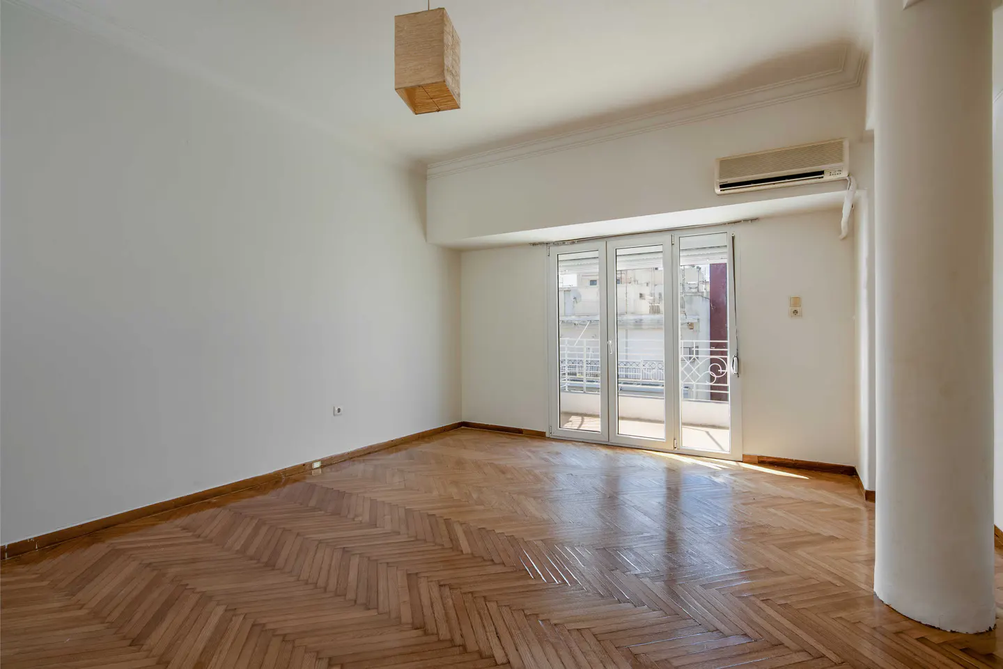 Bright, empty room with herringbone wood floors, white walls, and a balcony with white doors. A square lamp hangs from the ceiling.