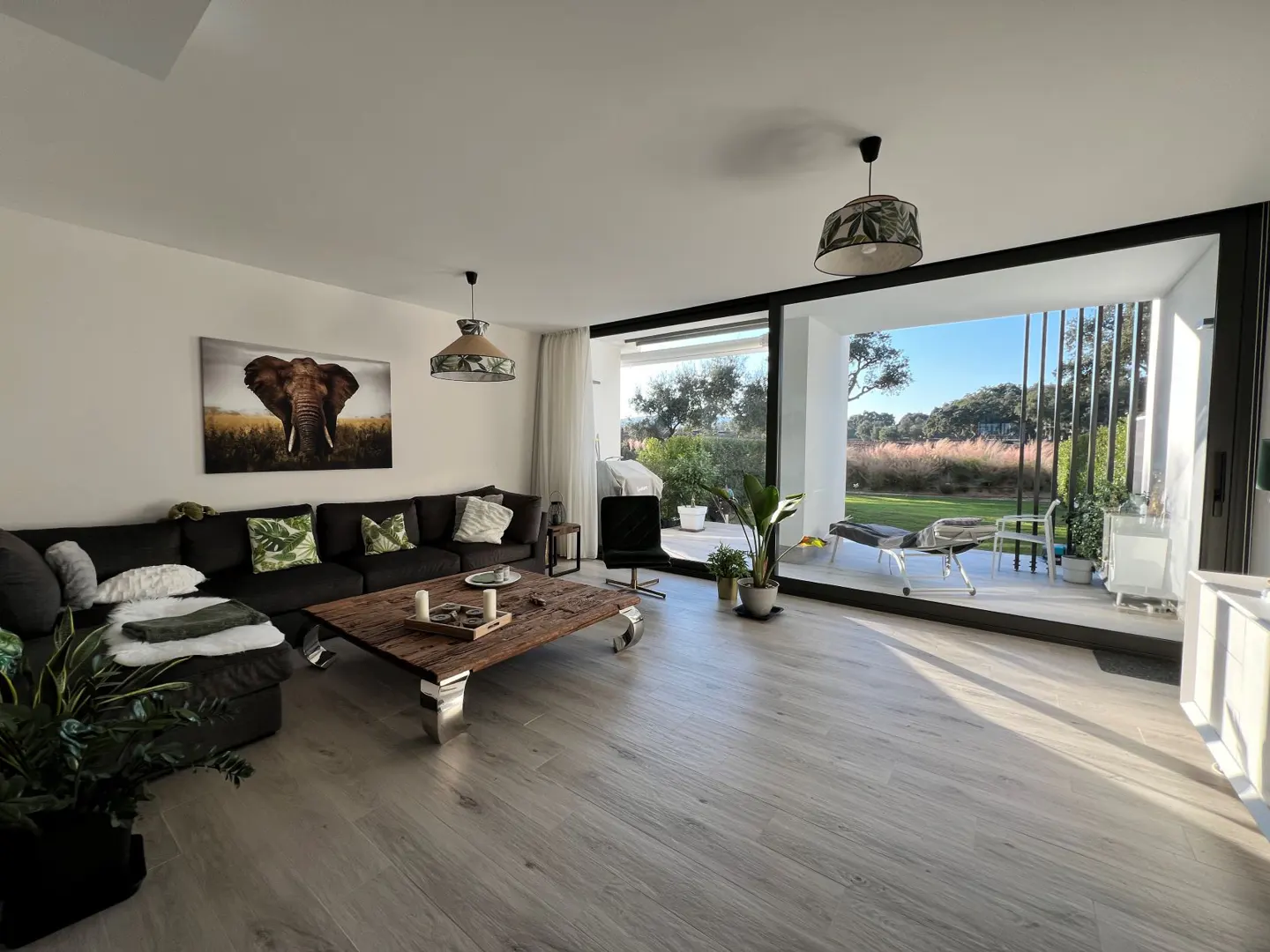 A bright living room with a large black sofa, wooden coffee table, and an elephant picture. Sliding glass doors open to a patio and garden.