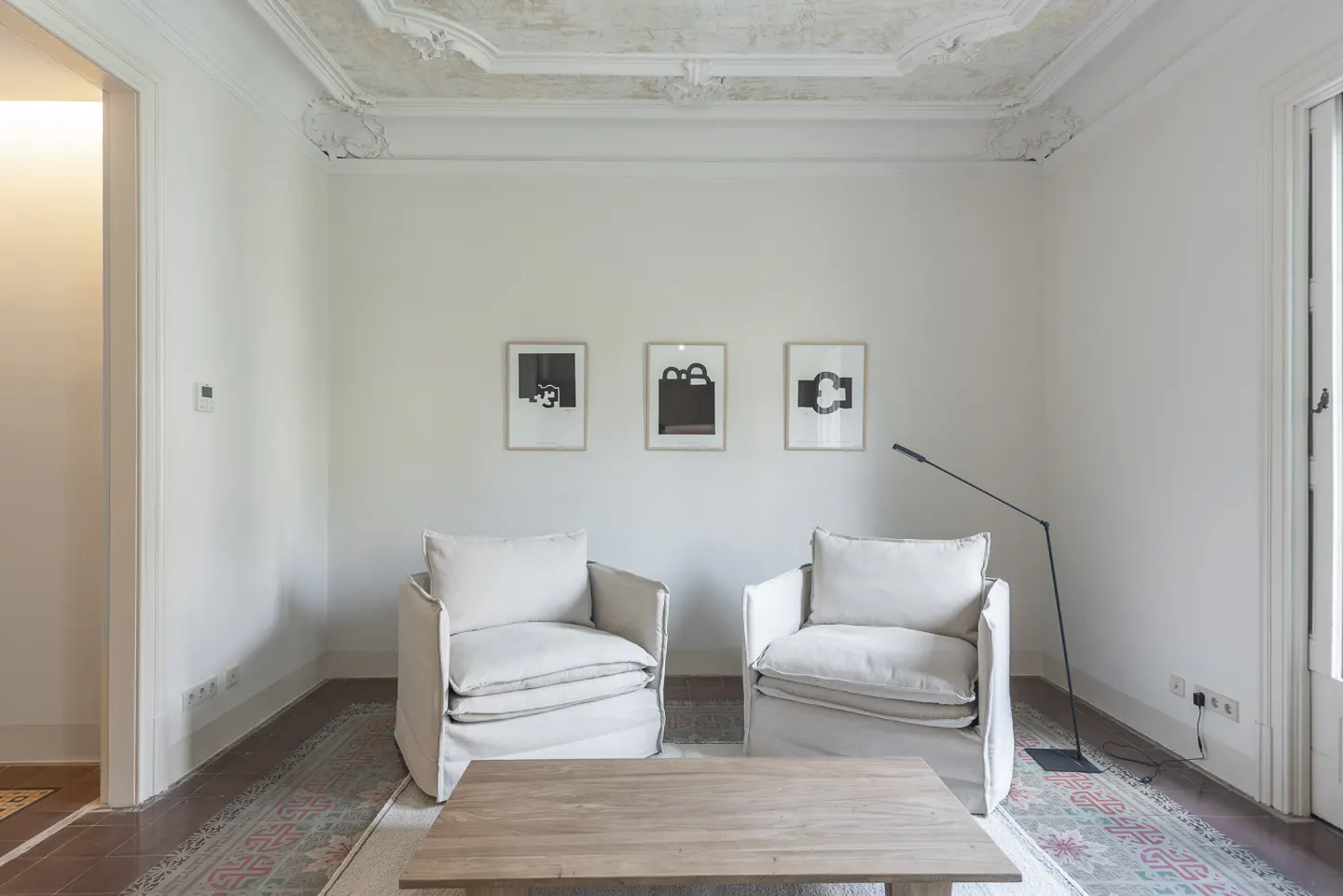 Bright living room with two white armchairs, a wooden coffee table, and three framed abstract art pieces on the wall.
