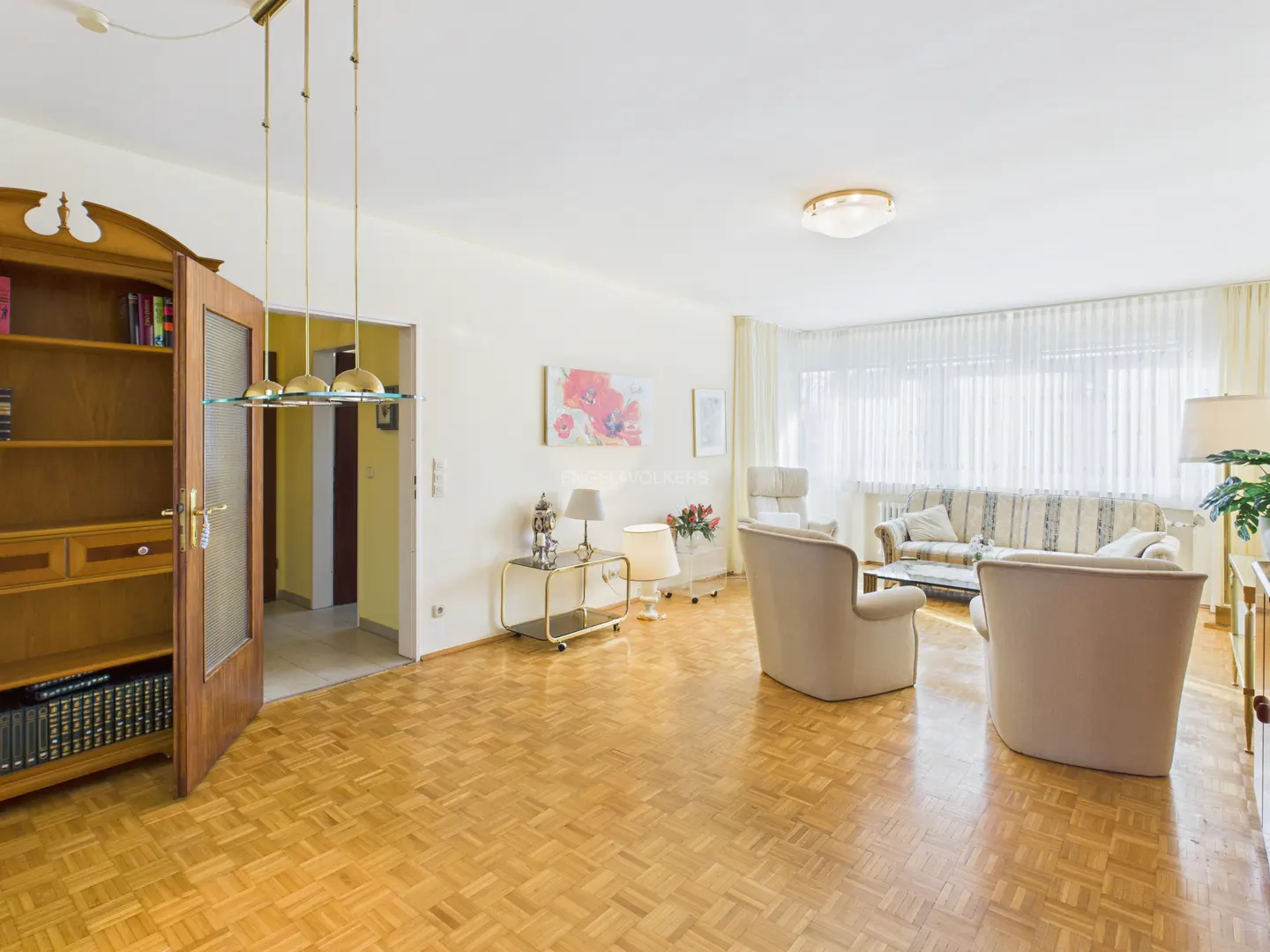 Bright living room with parquet floors, beige armchairs, and a striped sofa near sheer white curtains. A wooden bookcase stands to the left.