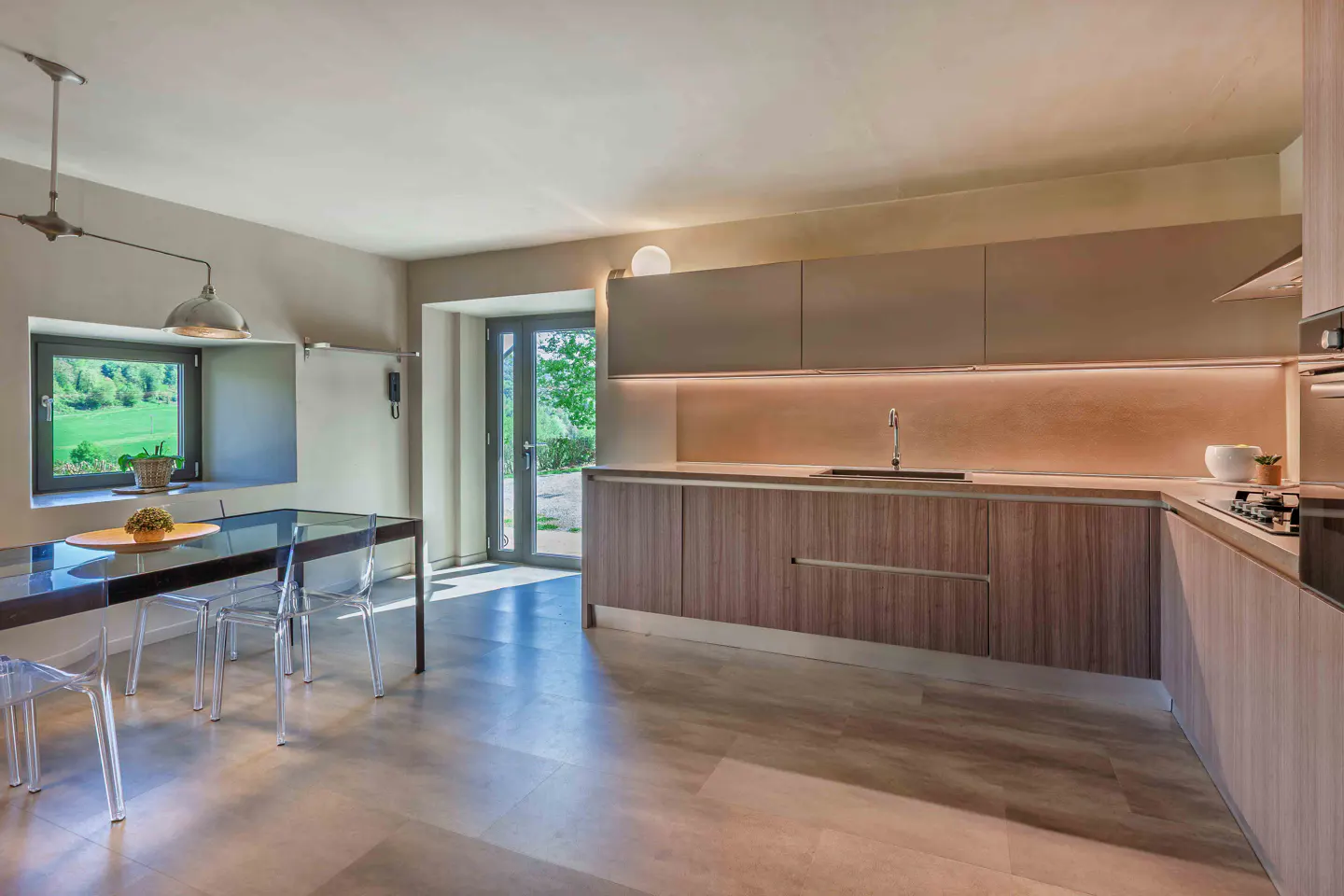 A modern kitchen with wood cabinets, a black table with clear chairs, and a window overlooking a green field.