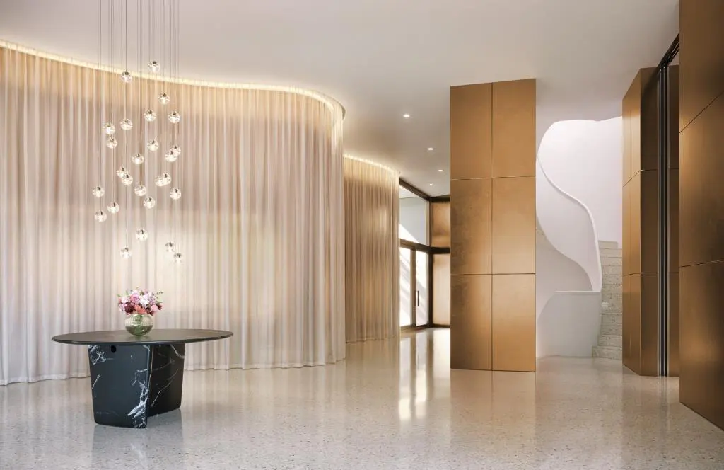 A modern lobby with a black marble table, floral arrangement, and a crystal chandelier. Gold panels and a curved curtain wall add elegance.
