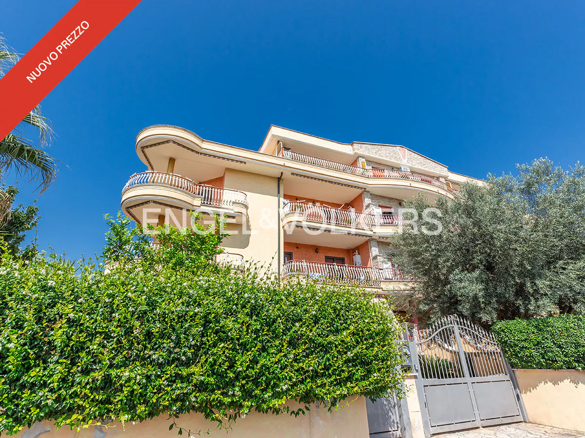 Three-story apartment building with balconies, green bushes, and a gray gate under a clear blue sky. "Nuovo Prezzo" banner.
