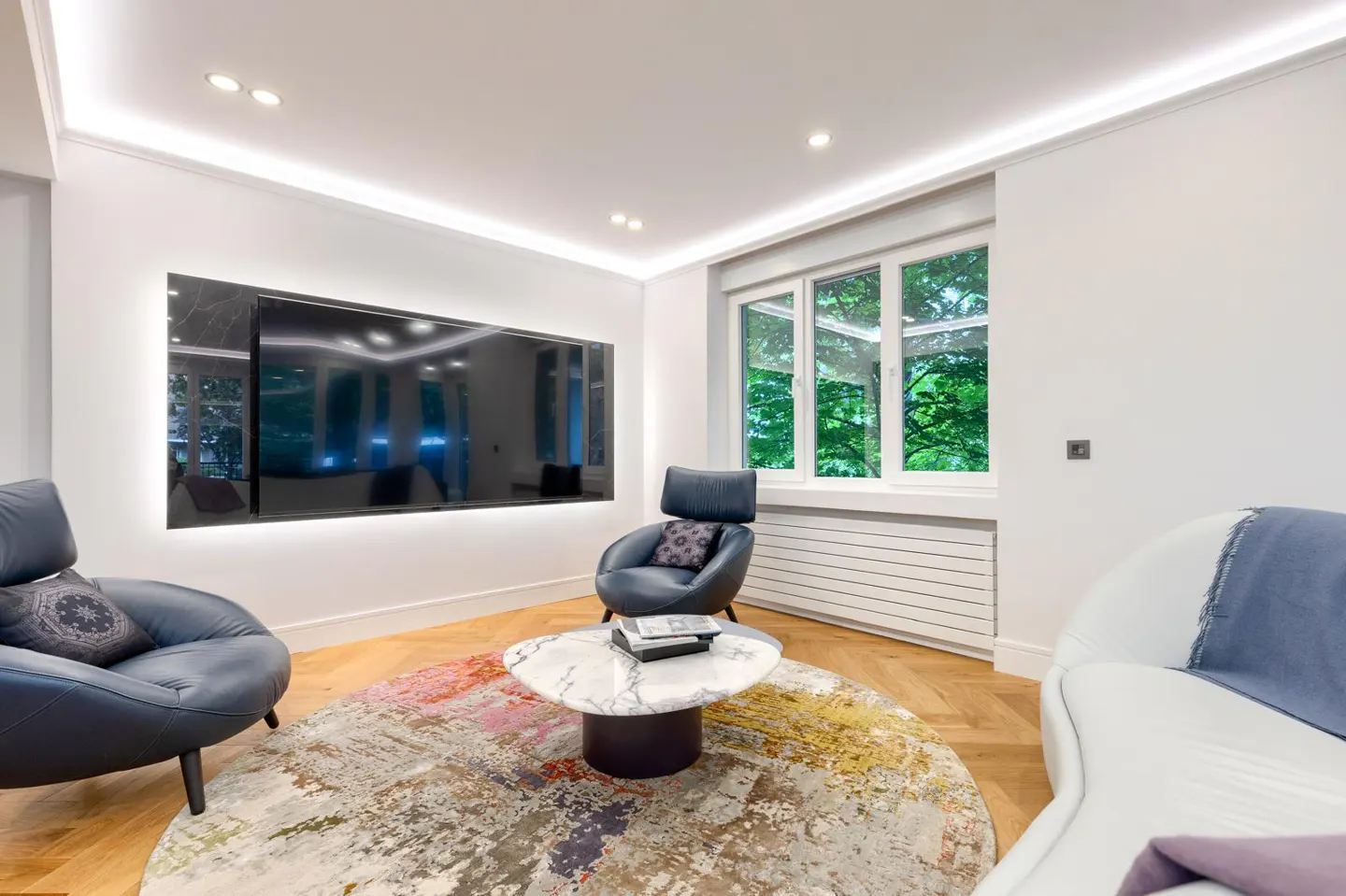 Bright living room with white walls, wood floors, and a large TV. Two blue chairs and a white sofa surround a marble coffee table on a colorful round rug. A window shows green trees.