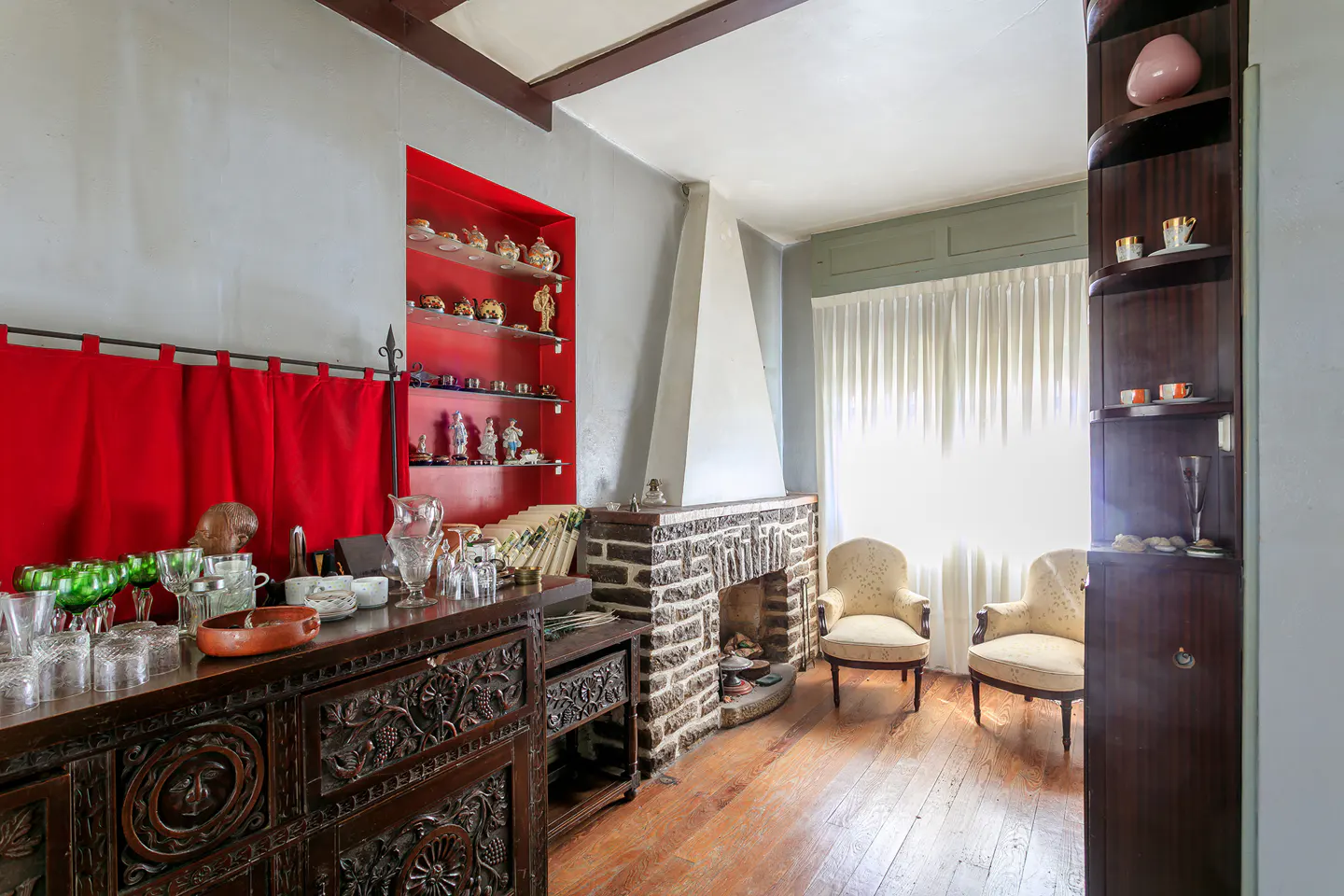 Living room with a dark wood carved cabinet, red shelves, stone fireplace, two chairs, and a dark wood corner shelf.