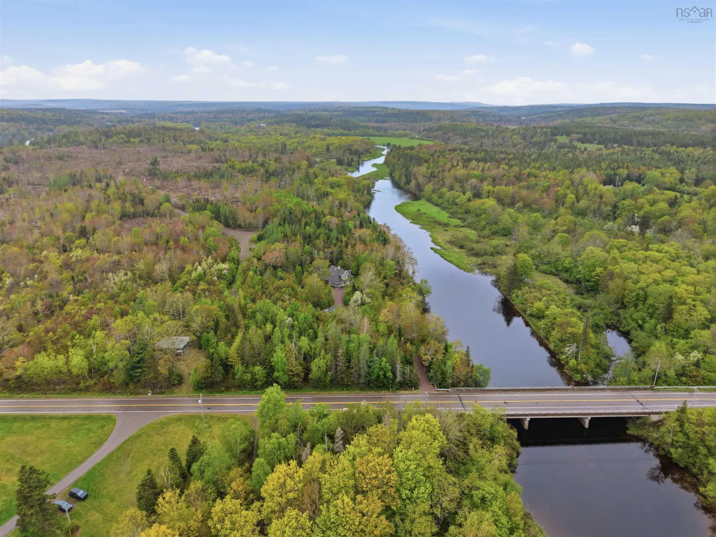 Aerial view of a river winding through a lush green forest, crossed by a road bridge, under a bright blue sky.