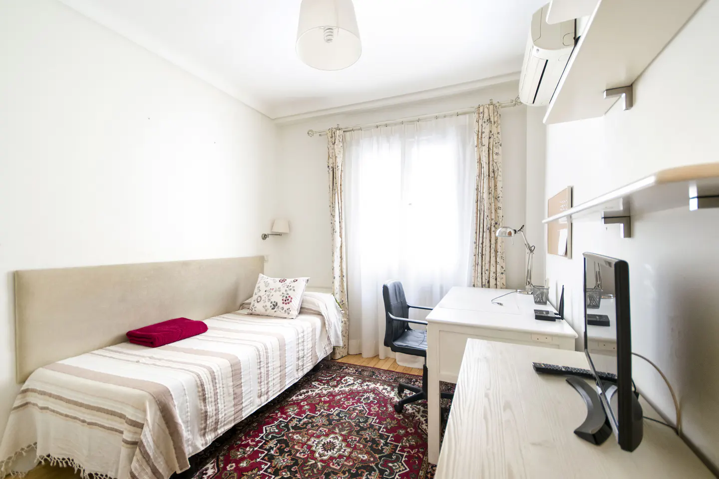 A bright bedroom with a single bed, desk, and red patterned rug.