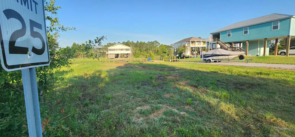 Vacant lot with a 25 MPH speed limit sign, green grass, and houses on stilts in the background.