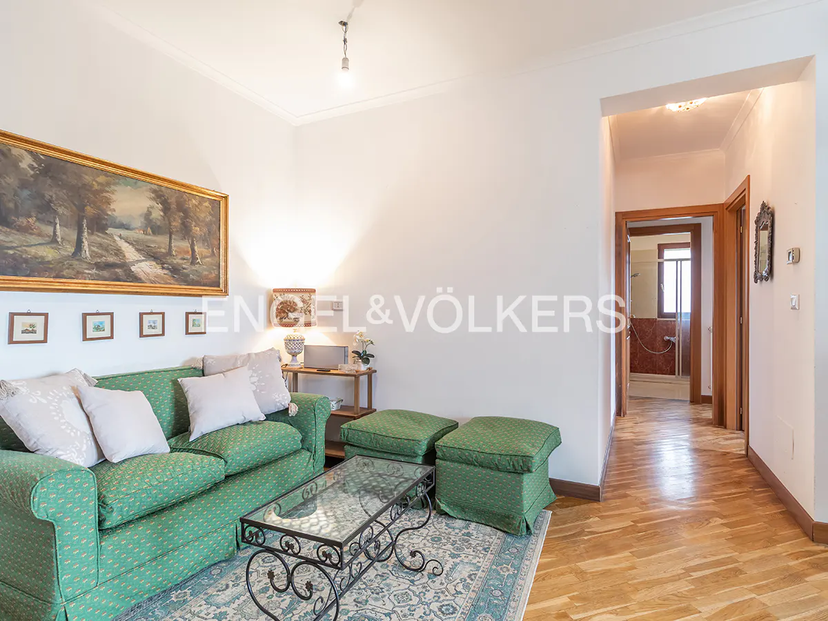 Living room with green sofa, ottomans, and glass coffee table on a patterned rug. A hallway leads to a bathroom. Artwork adorns the walls.