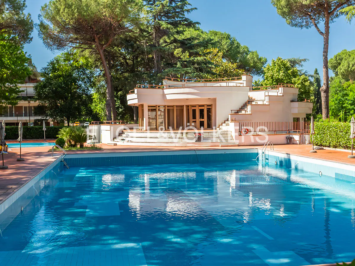 Large outdoor swimming pool with blue water in front of a modern white building surrounded by lush green trees.
