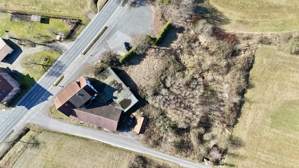 Aerial view of a building with brown roofs, a parking lot, and a wooded area next to a grassy field.