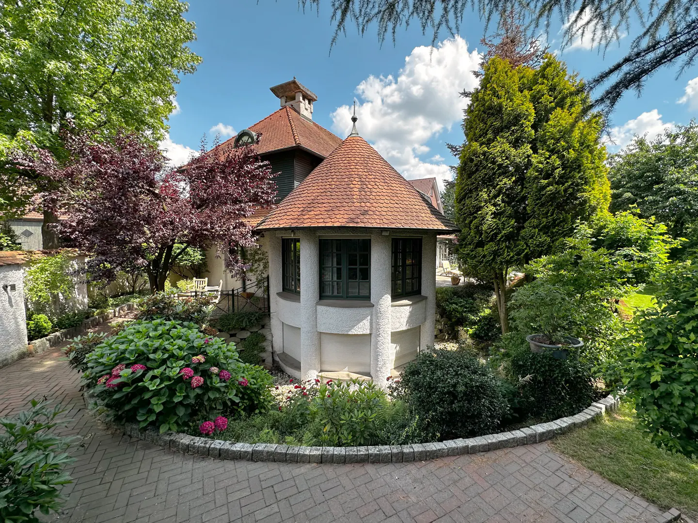 Exterior view of a white house with a round turret and red tile roof, surrounded by lush green trees and plants.