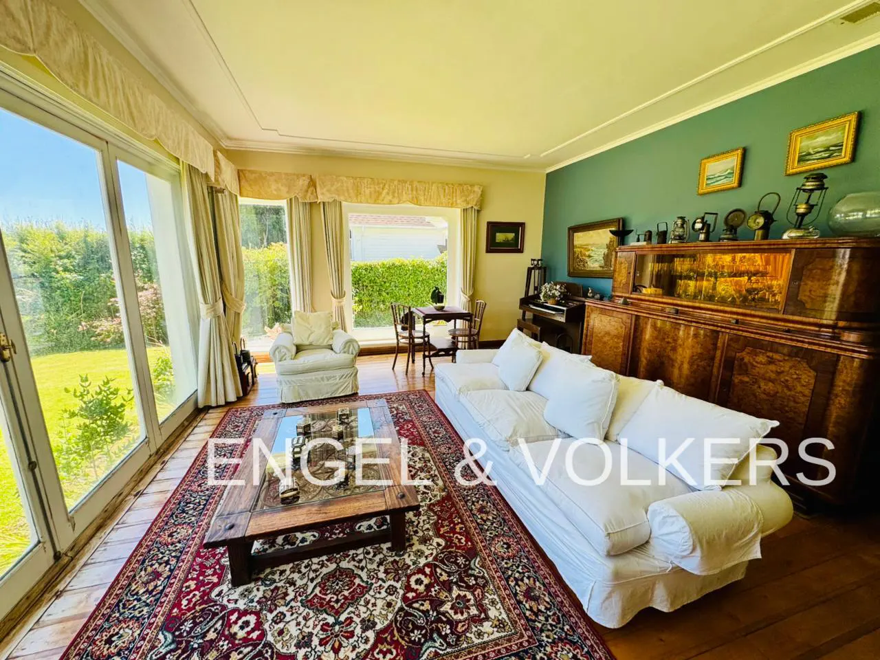 Bright living room with a white sofa, wooden coffee table on a patterned rug, and large windows overlooking a green garden.