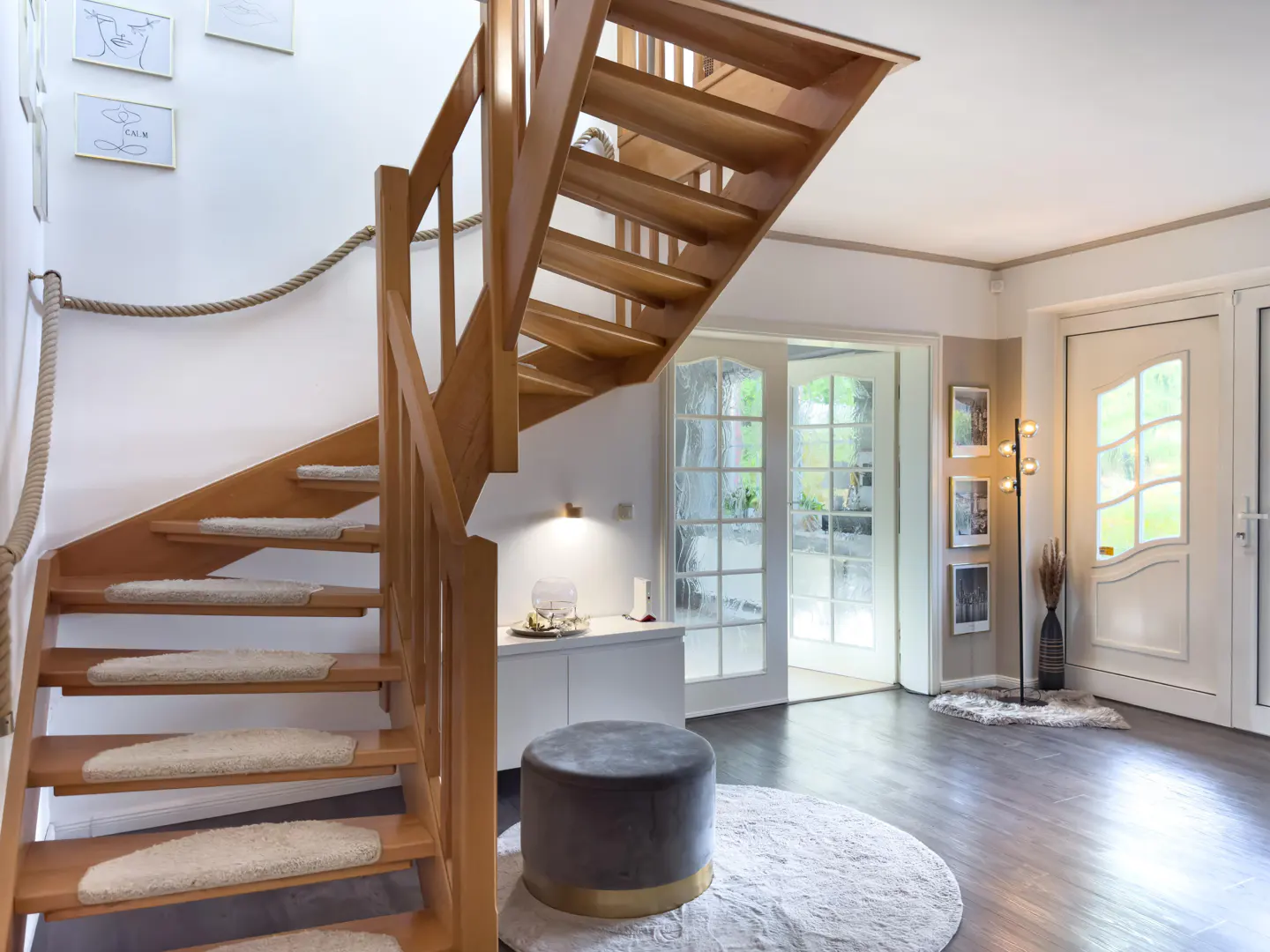 Bright foyer with wooden stairs, carpet treads, and rope railing. A gray ottoman sits on a white rug. Glass doors and a white door lead outside.