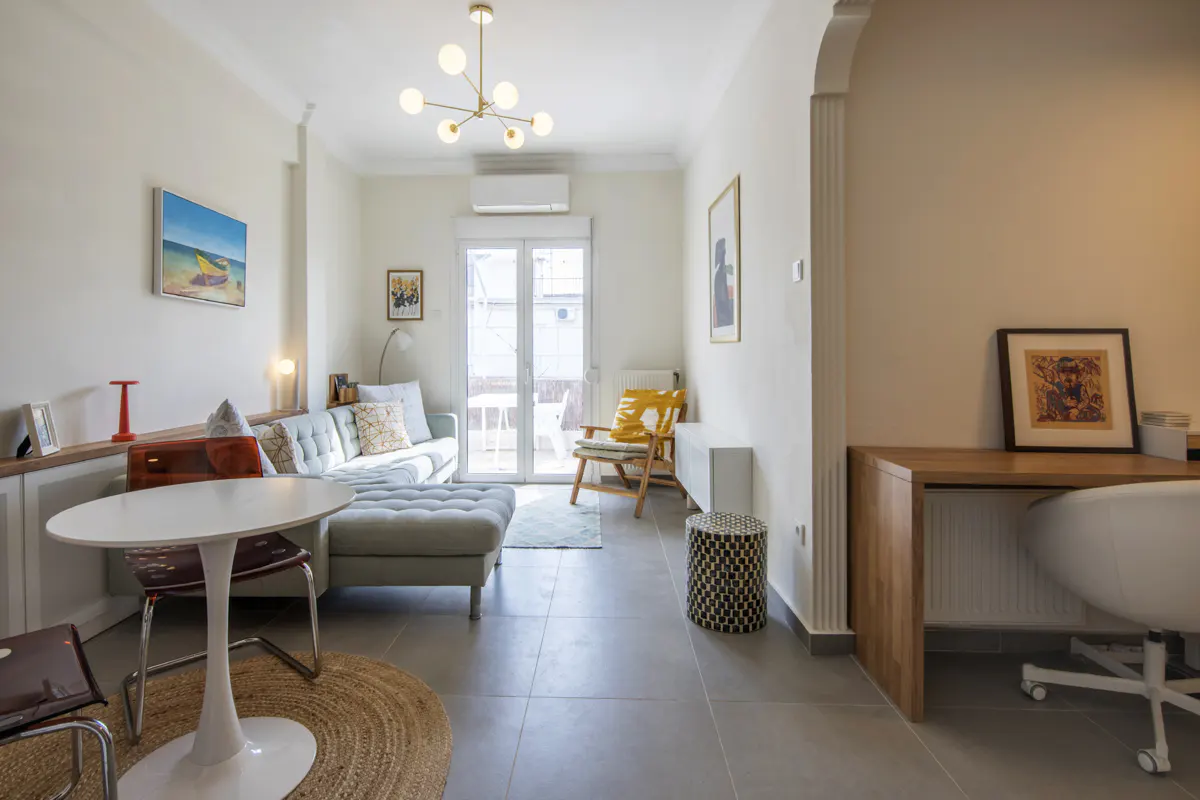 Bright living room with a gray sofa, white table, and a desk in the corner. Artwork adorns the walls, and natural light streams in through the balcony doors.
