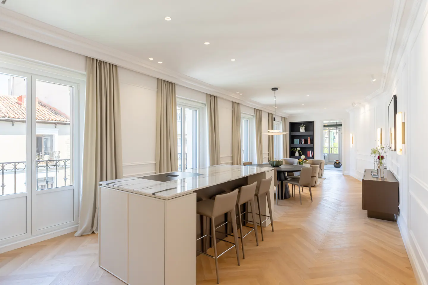 Modern open-plan kitchen and dining area with a marble island, bar stools, herringbone wood floors, and large windows.
