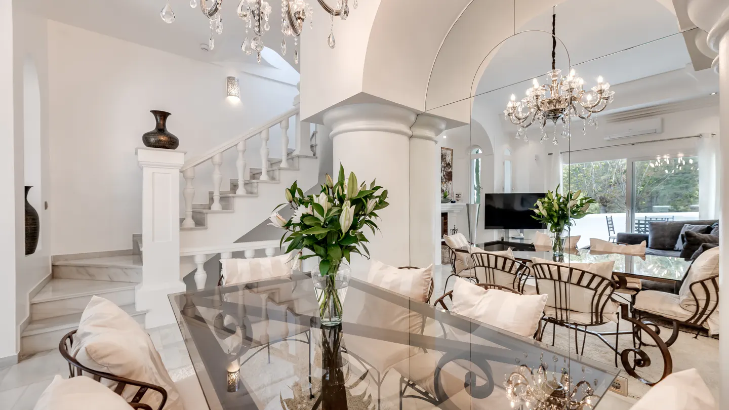 Bright, airy dining room with glass table, wrought iron chairs, white cushions, and lily centerpiece. Staircase and chandelier visible.