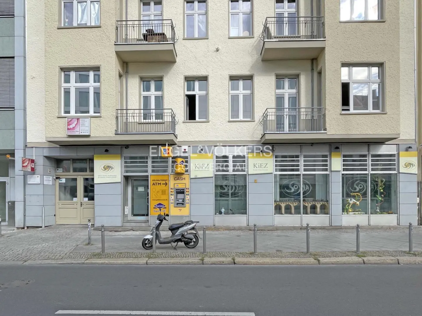 Exterior view of a light yellow building with balconies, an ATM, and a parked scooter on the sidewalk.